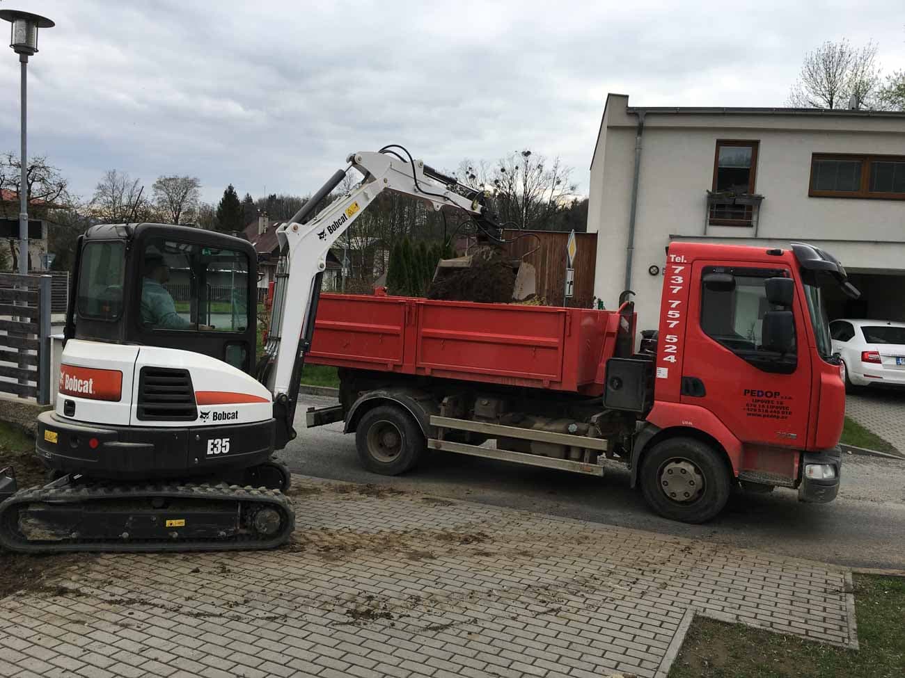 Minibagr Bobcat E35 nakládá zeminu na červené auto PEDOP u rodinného domu